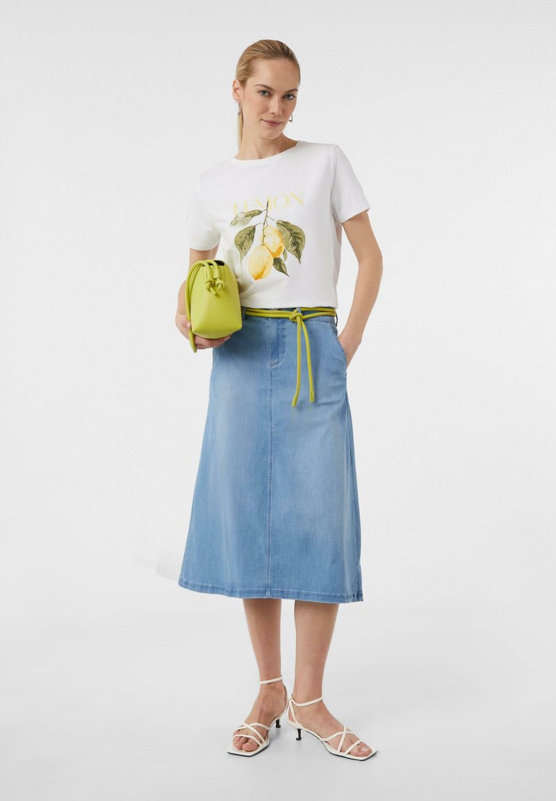 T-shirt graphique blanc avec imprimé citron, jupe midi en denim bleu, ceinture verte et petit sac vert. Modèle portant des talons blancs à lanières.