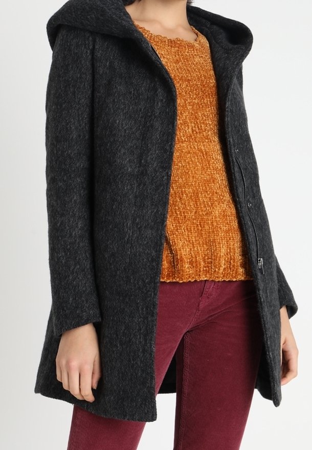 Personne portant un manteau à capuche gris foncé sur un pull orange texturé et un pantalon bordeaux, debout devant un fond blanc.