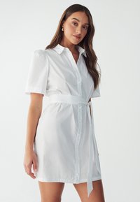 Valkoinen shirt-tyylinen mekko, jossa on lyhyet puffihihat, napit edessä ja matching-sidontavyö vyötäröllä. Sujuva puuvillakangas klassisella muotoilulla.