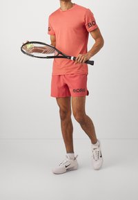 Korallfärgad performance t-shirt och shorts, med "BORG"-logotyp, håller en tennisracket med boll, och är ihop med vita Nike-skor.