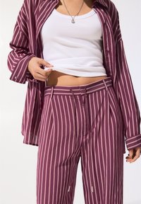 Set de cămașă și pantaloni cu dungi maro și roz, având o croială lejeră și închidere cu nasturi, purtat cu un top alb cu nervuri dedesubt.