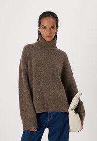 Maglione oversize marrone con collo alto realizzato in un materiale morbido e texturizzato; presenta maniche lunghe e larghe. Abbinato a jeans blu in denim.