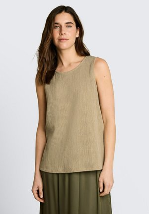 Vrouw met lang bruin haar, gekleed in een mouwloze, gestructureerde beige top en olijfgroene broek, staand tegen een effen lichte achtergrond.