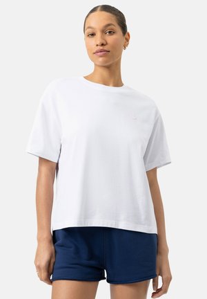 Junge Frau mit zurückgebundenem Haar, die ein lockeres weißes Kurzarmshirt und marineblaue Shorts vor einem einfarbigen Hintergrund trägt.