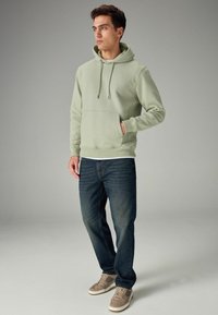 Sudadera con capucha verde claro y bolsillo canguro, combinada con jeans azul oscuro y zapatillas de color beige, sobre un fondo neutro.