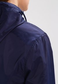 Navy blue hooded jacket made of lightweight, smooth fabric with visible stitching and adjustable drawstring on the hood.