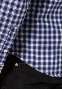 Blå och vit rutigt skjorta med knapp-ned krage och långa ärmar, tillverkad i bomull. Nederkanten är rundad och den är parat med svarta jeans.