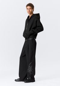 Μαύρο φούτερ με φερμουάρ και κουκούλα, συνδυασμένο με φαρδιά μαύρα παντελόνια που έχουν σχέδιο φλόγας από στρας στο αριστερό πόδι και μαύρα αθλητικά παπούτσια.