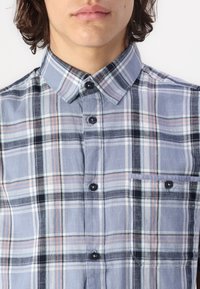 Lichtblauw geblokt shirt gemaakt van stof met een zachte textuur. Heeft een button-down kraag, zwarte knopen en een borstzak.