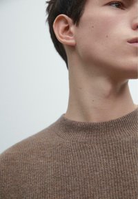 Brązowy prążkowany sweter z dzianiny z wysokim kołnierzem, o fakturowanym wykończeniu i subtelnych cętkach na całej powierzchni, noszony na jednolitym tle.