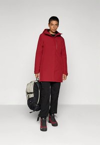 Raudonas neperšlampamas paltas su gaubtu, priekyje užtrauktukas, derinamas su juodomis kelnėmis ir batais. Nešioja pilką ir juodą kuprinę.