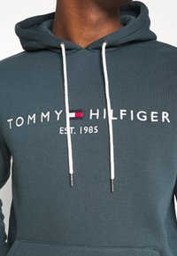 Sudadera con capucha en verde azulado oscuro, con cordones blancos, logotipo bordado "TOMMY HILFIGER" y detalle de "est. 1985" en hilo blanco.