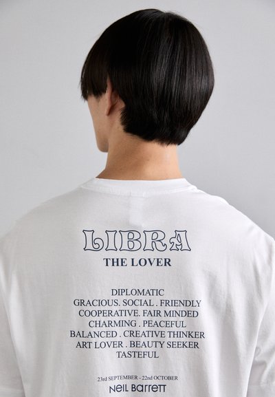 Baltas medvilnės marškinėliai su mėlyna spalva užrašytomis „LIBRA“ ir „THE LOVER“ frazėmis. Papildomi aprašomieji žodžiai atspausti žemiau, sukrauti vienas ant kito.