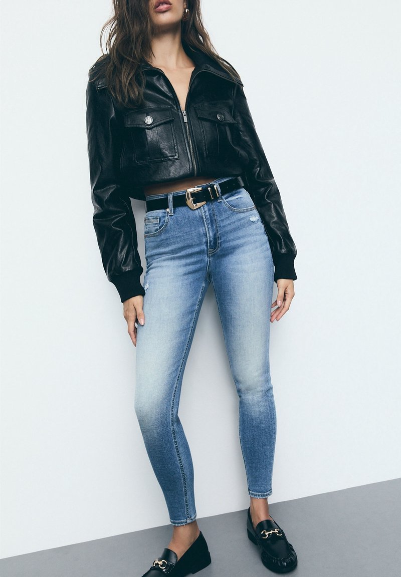 Chaqueta corta de cuero negro con dos bolsillos frontales, combinada con jeans ajustados de color azul claro y mocasines negros. Un cinturón simple acentúa la cintura.