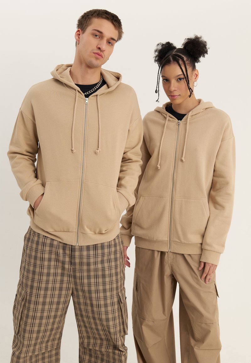 Two young adults wearing beige zip-up hoodies and casual pants, standing against a plain light background.