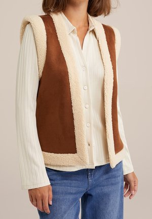 Femme portant un gilet en peau lainée marron sans manches par-dessus une chemise blanche côtelée boutonnée et un jean bleu, debout les mains détendues.