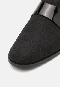 Zapato de tela negra con una correa lisa y brillante negra en la parte superior. Punta redondeada y suela de goma con una ligera textura.