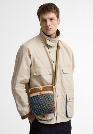 Νέος άνδρας που φοράει μπεζ μπουφάν με ψηλό γιακά και καφέ τσάντα ώμου με μπλε δίχτυ στην πρόσοψη, στέκεται μπροστά σε λευκό φόντο.