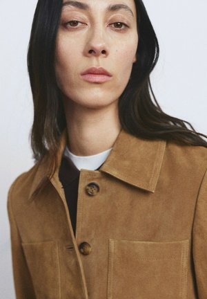 Mujer con cabello largo y oscuro, vistiendo una chaqueta de ante color marrón claro con botones al frente y un gran cuello, sobre un fondo claro y liso.