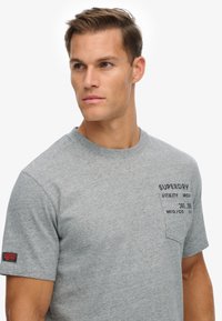 Camiseta de algodón gris de manga corta con un logotipo impreso en el bolsillo del pecho y una etiqueta de tela en la manga izquierda. Textura suave.