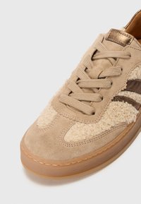 Baskets beiges avec des accents en daim et en textile moelleux, bout rond, fermeture à lacets sur le devant, et une semelle en caoutchouc translucide, avec un détail métallique.