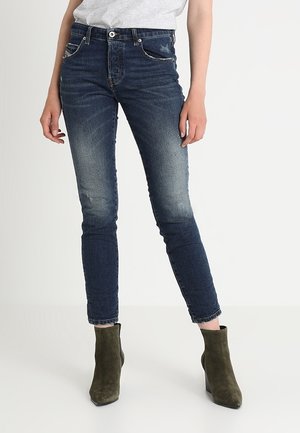 Kvinde iført falmede mørkeblå skinny jeans og olivengrønne, ruskindede ankelstøvler, stående mod en ensfarvet lys baggrund.