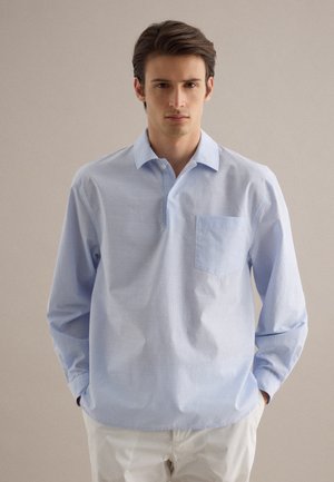 Mann mit braunen Haaren, der ein hellblaues, langärmeliges Hemd mit Kragen und Brusttasche trägt, die Hände in den Taschen weißer Hose, neutraler Hintergrund.