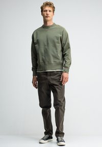 Olijfgroene sweatshirt met een ronde halslijn en rib gebreide mouwen, gecombineerd met donkerbruine broeken en zwarte sneakers met witte zolen.