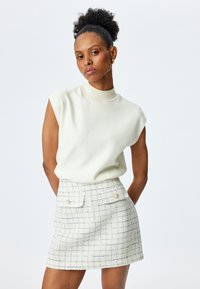 Top sin mangas de color crema claro con cuello alto, combinado con una falda mini ajustada y texturizada de estampado de cuadros que cuenta con dos bolsillos frontales.