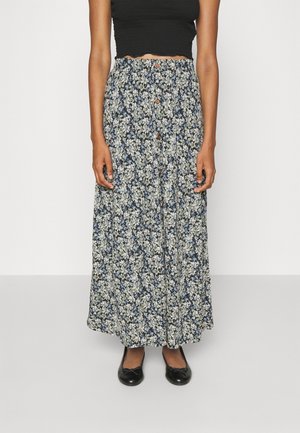 Kvinde iført en sort cropped top, høj-taljet lang blomsterprintet nederdel med knapper foran og sorte balletsko, stående foran en ensfarvet baggrund.