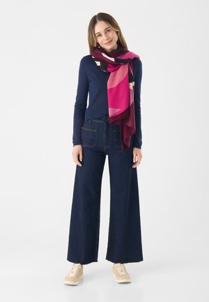 Donkerblauwe trui met lange mouwen en V-hals, gecombineerd met brede denim jeans in een donkere kleur en een kleurrijke geometrische sjaal in roze en bordeaux.