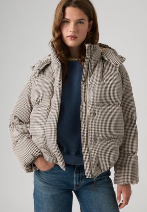 Jeune femme portant une doudoune beige et noire à carreaux par-dessus un sweat-shirt bleu et un jean, debout devant un fond clair.