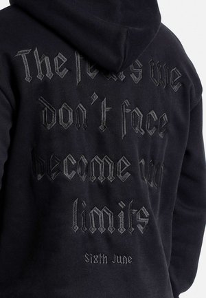 Schwarzer Kapuzenpullover mit strukturiertem, dunkelgrauem gesticktem Text, der besagt: "Die Ängste, denen wir uns nicht stellen, werden zu unseren Grenzen" mit "Sixth June" darunter.
