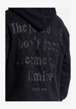 Sudadera negra con texto bordado texturizado en gris oscuro que dice: "Los miedos que no enfrentamos se convierten en nuestros límites" y "Sixth June" debajo.