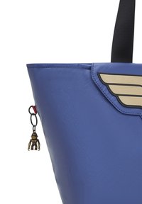 Blå, glansig tote-väska med en guldvingedesign på locket och en liten apcharm fäst med en röd slinga. Slät textur, strukturerad form.
