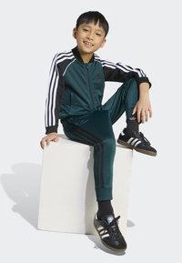 Teal jumpsuit med svarta ränder och manschetter, buren av ett barn som sitter på en vit kub. Svarta sneakers med vita detaljer.