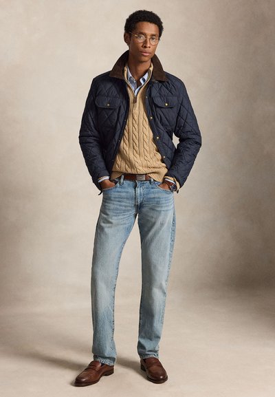 Mand iført briller, navy blå quiltet jakke, tan kabelstrikket sweater, lyseblå jeans og brune lædersko, stående med hænderne i lommerne.