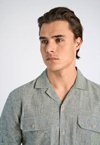 Camisa de manga corta, con cuello, en lino verde claro con un acabado texturizado. Presenta dos bolsillos en el pecho y botones en tono marrón.