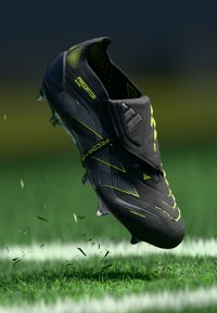 Black football boot with yellow accents, textured upper, and prominent grooves. It features a firm heel, lightweight construction, and visible studs.