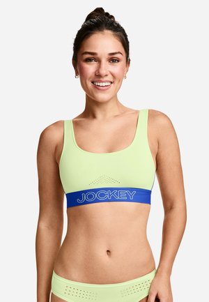 Lächelnde Frau mit Dutt, die einen hellgrünen Sport-BH und passende Unterwäsche mit einem blauen Jockey-Logo-Band am BH trägt.