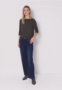 Žena stojící s dlouhými blond vlasy, oblečená v černém tričku s dlouhým rukávem, tmavě modrých širokých džínách a hnědých kotníkových botách na jednobarevném pozadí.