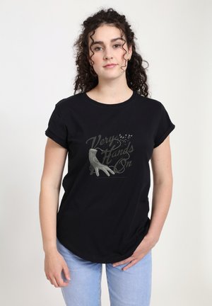 Woman with curly hair wearing a black T-shirt with a vintage glove and "Very Hands On" text, paired with light blue jeans, standing against white background.