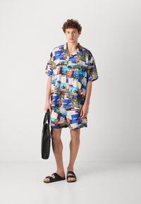 Kortærmet skjorte med krave og matchende shorts i en collage af farverige postkorttryk; båret med sorte sandaler og en strandtaske.