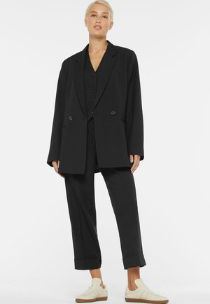 Blonde Frau steht, trägt einen übergroßen schwarzen Blazer, schwarze Hosen mit hochgekrempelten Bündchen, eine schwarze Weste und weiße Turnschuhe vor weißem Hintergrund.