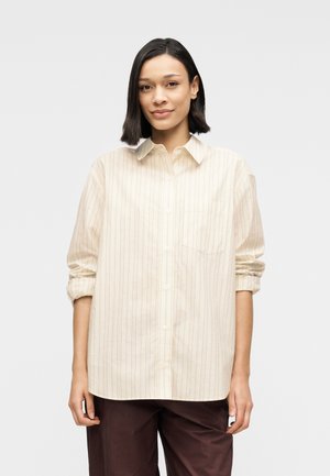 Chemise beige rayée à boutons avec col et une poche poitrine unique. Le tissu est léger avec des manches retroussées. Portée avec un pantalon marron foncé.