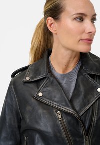 Svart lädermotorcykeljacka med silverdetaljer, sned dragkedja och breda slag; bärs över en grå tröja. Strukturerad yta.