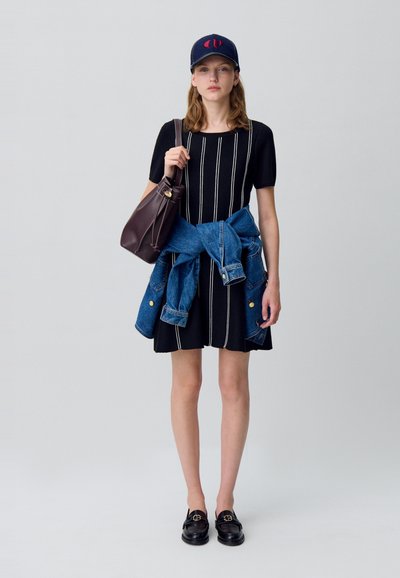 Mujer joven con vestido negro de rayas blancas verticales, chaqueta de denim atada a la cintura, gorra de béisbol, mocasines negros y llevando un bolso marrón.