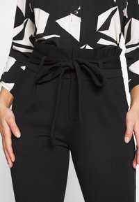 Svarta högmidjade byxor med en tygrem knut, som har en slät textur och sidofickor. I kombination med en mönstrad blus.