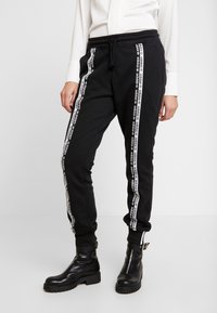 Svarta sweatpants med vita sidostråk som har upprepade logotext. Gjorda av mjukt material, med elastisk midja och ärmslut.