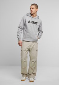 Felpa grigia con la scritta "ARMY" stampata in nero, abbinata a pantaloni cargo beige con tasche laterali. Il modello indossa sneaker bianche con suole gialle.